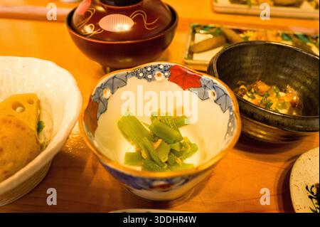 Lightly seasoned green mountain vegetables are served in a decorative porcelain bowl as part of a traditional Japanese breakfast spread. Warm lighting highlights the textures and vibrant colors of the assorted dishes on a wooden table. Stock Photo