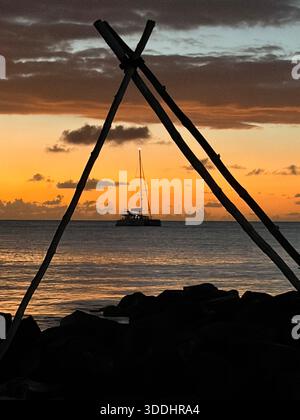 Sailing yacht anchored at sunset, framed by a traditional wooden shoreline structure in Antigua, Caribbean Sea. Stock Photo