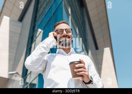 Handsome mature man manager with coffee to go having business call, standing against modern office building Stock Photo