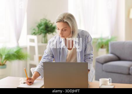 Focused Elderly Businesswoman Working On Laptop And Notepad In Home Office Stock Photo