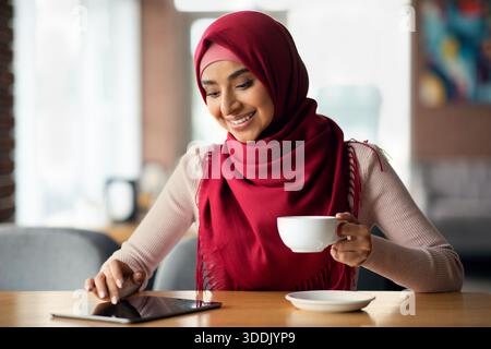 Woman in hijab chilling at cafe alone, using digital tablet Stock Photo