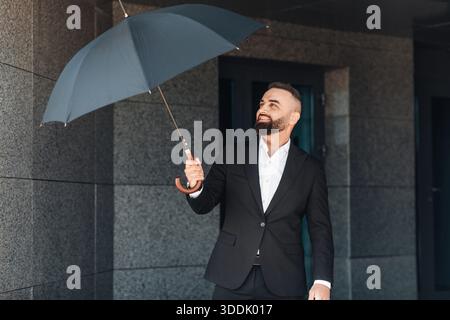Mature office worker wearing classical suit holding umbrella while walking outdoors, going home after work in office Stock Photo