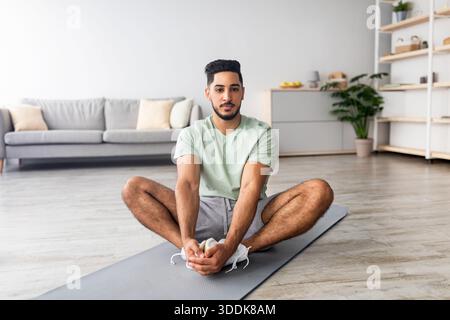 Home workout. Sporty young Arab guy stretching on mat in living room, practicing yoga, full length Stock Photo