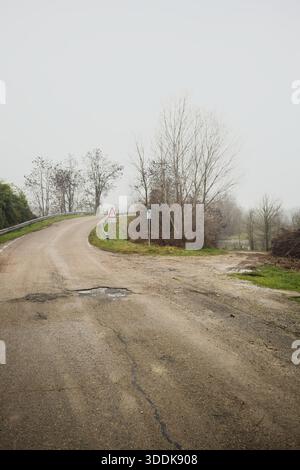 Fork between a road on an embankment, leading to a bridge, and a muddy trail, leading to a river framed by trees, on a misty and cloudy day in the ita Stock Photo