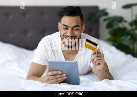 Young Middle Eastern Man Enjoys Shopping Online While Relaxing in Bed at Home Stock Photo