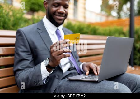Young african american businessman using credit card and laptop, doing some online shopping, sitting outdoors Stock Photo