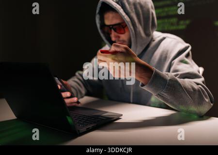 Man in hoodie holding phone and credit card, performing cybercrime, phishing and online scam operations, surrounded by green digital code on dark Stock Photo