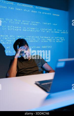 Vertical portrait of confused young male programmer facing challenging coding problem, feeling frustrated while working on laptop with lines of blue Stock Photo