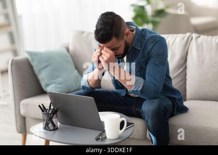 Stressed young Arab man sitting on couch near laptop, having problem with online work or studies at home Stock Photo
