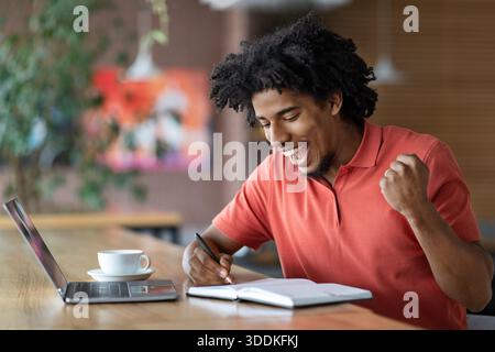 Glad happy excited curly handsome young black man make notes in notebook, victory gesture in cafe interior Stock Photo