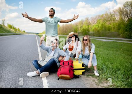 Unhappy millennial tourists sitting on roadside, cannot catch car, waiting for too long, having no smartphone connection Stock Photo