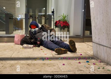 A person sleeps on a paved ground, wrapped in blankets. Scattered confetti and a drink bottle lie nearby. Stock Photo