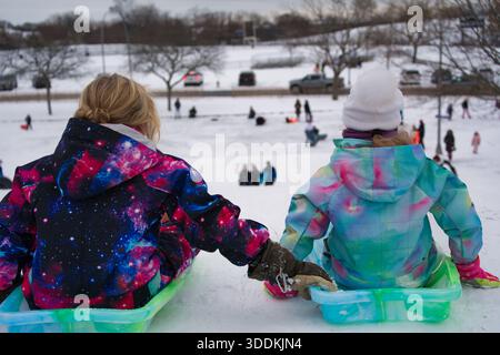 Two children in brightly colored snowsuits sit on sleds, holding hands, looking down a snow-covered hill with other people playing. Stock Photo