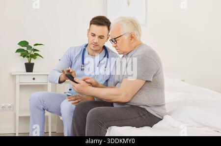 Young male caregiver teaching senior person to handle with mobile phone sitting on bed in clinic. Stock Photo