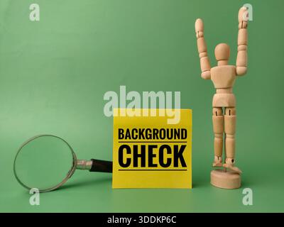 Yellow sticky note with the text BACKGROUND CHECK on a green background, accompanied by a magnifying glass and a wooden artist's figure. Stock Photo