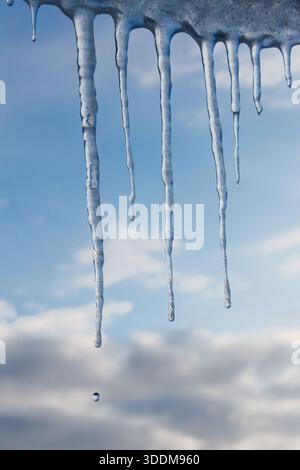 Transparent icicles hanging from roof edge with single water drop falling, soft blue sky and clouds in background, concept of thaw, changing seasons a Stock Photo