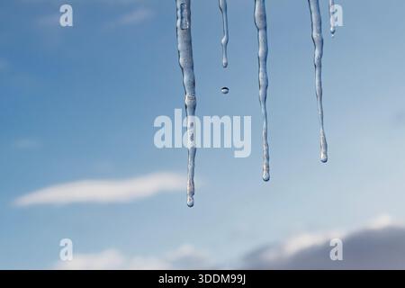 Transparent icicles hanging from roof edge with single water drop falling, soft blue sky and clouds in background, concept of thaw, changing seasons a Stock Photo