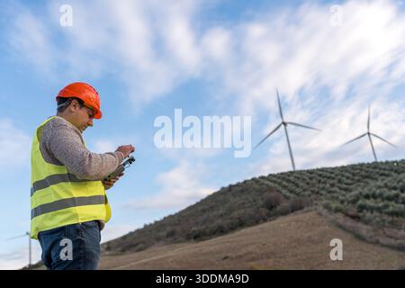 Engineer wearing safety vest and hard hat operating a drone controller for inspection and maintenance at a wind farm Stock Photo