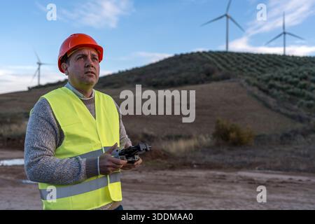 Worker is seen controlling a drone while monitoring wind turbines on a hill, dressed in a safety vest and hard hat Stock Photo