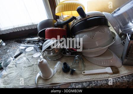 Kitchen countertop showing a large stack of clean, wet dishes, pots, pans, and utensils drying on a towel after washing, representing household chores Stock Photo