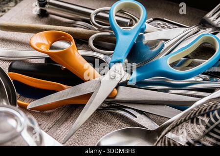 Kitchen drawer showing colorful plastic handled scissors mixing with various stainless steel cutlery, including spoons, forks, and other utensils crea Stock Photo