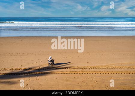 A lone person sits in a chair on smooth golden sand, facing the rolling ocean. The expansive horizon and calm waves convey solitude, peace, and a mome Stock Photo