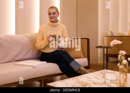 Woman sits on couch with a mug in a cozy indoor space Stock Photo
