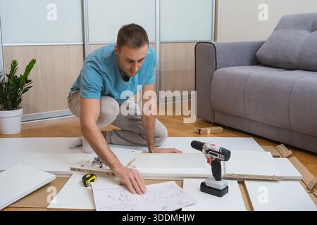 man sits on the floor reading furniture assembly instructions with tools and flat-pack parts scattered around him in a bright, modern living room Stock Photo