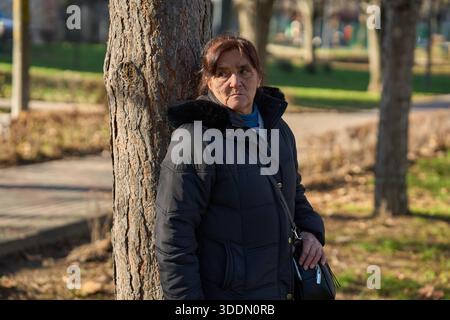 Senior woman leaning against a tree trunk in an autumn park with a serious thoughtful expression Stock Photo