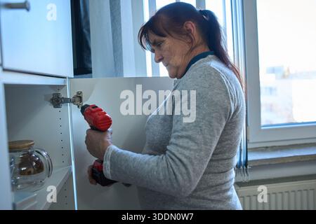 Senior woman using a cordless drill to fix a cabinet door hinge in her kitchen at home Stock Photo