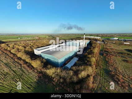 Aerial view of an industrial building with a tall chimney releasing dark smoke into the clear blue sky, surrounded by green fields, ELY, England, Unit Stock Photo