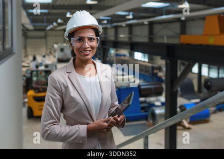 Young Indian woman, a successful metalworking factory manager writing notes after production inspection, smiling portrait shot. Stock Photo