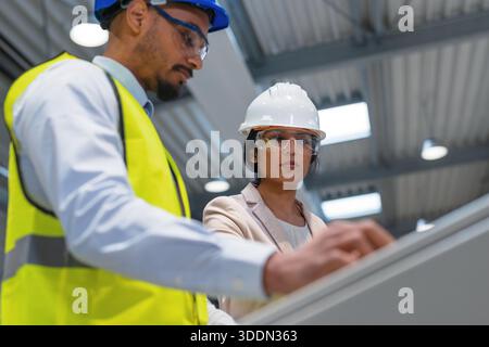 Indian female factory boss training African American male worker, in blue helmet and yellow vest, to work on manufacturing machinery. Stock Photo