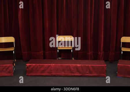 Indoor stage setup with red velvet curtains and wooden chairs arranged for an event. Stock Photo