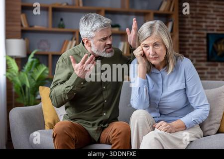 Mature man loudly gesturing and yelling at his upset wife, who is sitting on the couch and looking down, feeling sad and distressed about their relationship problems Stock Photo