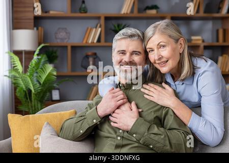 Happy senior couple smiling and experiencing domestic bliss while embracing on a comfortable sofa, representing love, companionship, and a fulfilling life after retirement Stock Photo