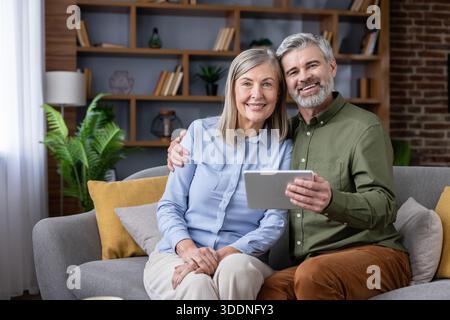 Smiling mature couple embracing on the living room sofa, happily using a digital tablet together, connecting with technology and enjoying their comfortable retirement lifestyle Stock Photo