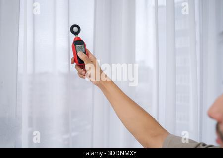 Close-up cropped shot of man measuring natural light near window with digital luxmeter, testing illumination levels for optimal home brightness Stock Photo