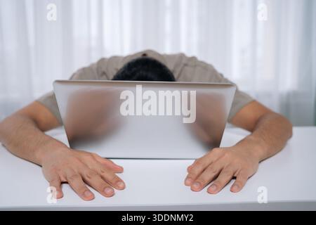 Exhausted man feeling burnout and stress, laying sleeping with face on laptop after working long hours, experiencing fatigue and overwork in workplace Stock Photo