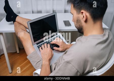 Young man working remotely on laptop in cozy home office, feet up on desk, casual t-shirt and shorts, focused and relaxed while handling freelance Stock Photo