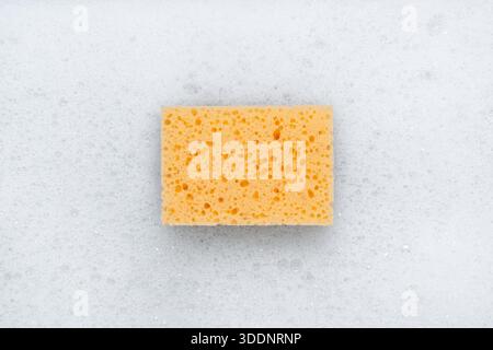 Yellow sponge on a white soapy foam background. Top view.м Stock Photo