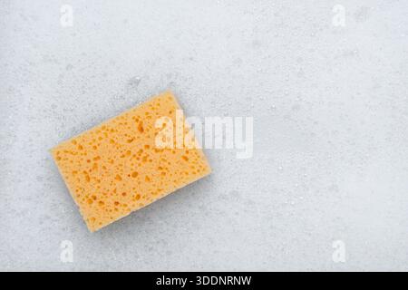 Yellow sponge on a white soapy foam background. Top view.м Stock Photo