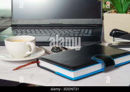 Blue Notebook Beside Laptop Coffee Cup On Tidy Desk With Glasses And Pen, Smartphone And Potted Plant Near Laptop, Soft Morning Light Creating Warm Pr Stock Photo