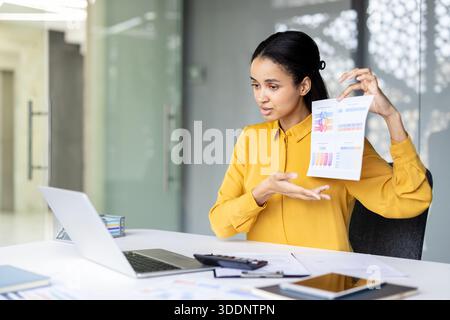 Businesswoman discussing charts and graphs on a printed report while engaging in a video conference from her modern office, analyzing data and sharing information remotely Stock Photo