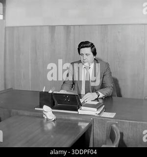 Slaviansk, Ukrainian SSR, Soviet Union, 1980s. An authentic archival working portrait of Comrade Novikov, director of the AIZ Armature and Insulator Plant, sitting at his desk in a wood-paneled office. The man is dressed in a suit and tie, focused on reading documents and handling business papers. The desk features a calendar stand, books and a decorative ceramic shoe. This vintage black and white photograph captures the daily routine of a Soviet industrial leader and the stability of the peaceful Donbas region during the USSR era. Stock Photo