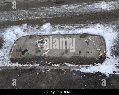 Weathered Concrete Cover On Snowy Roadway, Showing Chipped Paint, Rust Stains, Salt Residue And Granular Grit Along Asphalt Edge, Evoking Gritty Urban Stock Photo