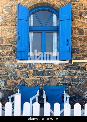 Arched window with vibrant blue shutters on a rustic stone wall, with two chairs and a white fence Stock Photo