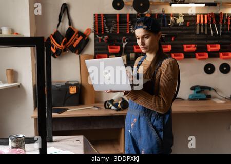 Woman carpenter in blue overalls typing laptop in workshop. worker artist using app online, Female craftsman Stock Photo