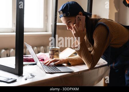 Female carpenter in blue overalls working in her workshop talking on smartphone taking order and using laptop computer at workplace Stock Photo