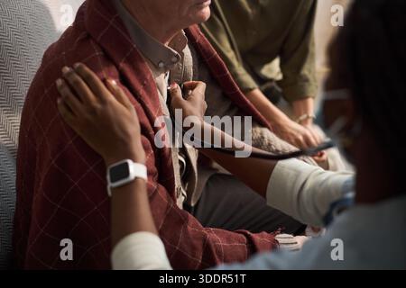 Middle aged Black woman using stethoscope examining chest of senior Caucasian man sitting on couch while another observing, hands and partial faces visible, focus on medical care interaction Stock Photo